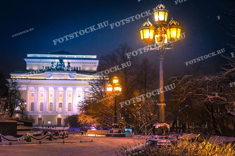Фотография показывает Александринский театр в Санкт-Петербурге, освещённый в зимний вечер. Тёплый свет уличных фонарей создаёт уютную атмосферу на фоне снежного парка. Идеально подходит для использования в культурных публикациях и туристических материалах.